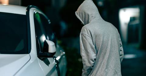 auto-inbraken; man met grijze hoodie aan en capuchon op loopt in het donker langs een witte auto en kijkt naar binnen 